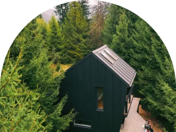 Een luchtfoto van een modern, zwart houten natuurhuisje met een puntdak, diep verscholen tussen de groene dennenbomen