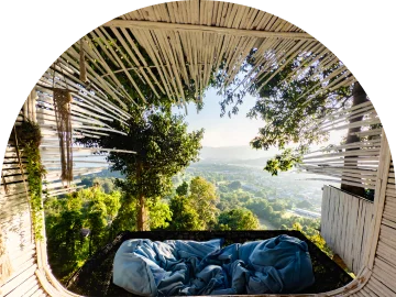 Een comfortabel buitenbed met blauwe kussens en dekens op een houten terras, met een panoramisch uitzicht op een groene vallei onder een warme zon