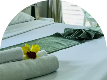 Een netjes opgemaakt hotelbed met witte lakens, een groene bedloper en zorgvuldig opgerolde handdoeken versierd met een gele orchidee