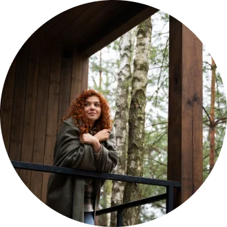 Een rond portret van een jonge vrouw met krullend rood haar, warm ingepakt in een bruine deken, leunend tegen de reling van een houten balkon in het bos