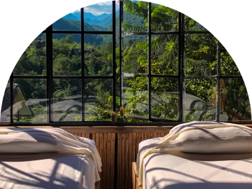 Uitzicht vanuit een slaapkamer met twee bedden door grote, klassieke ruitjesramen die uitkijken op een dichtbegroeid groen bos onder de zon